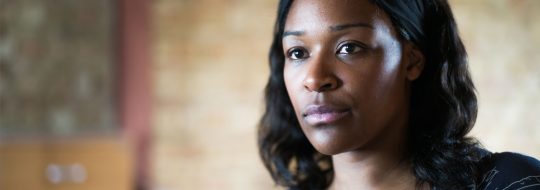 Charlene James, a black woman, looking off to something in her distance. She has shoulder length wavy black hair, dark brown eyes and closed lips.